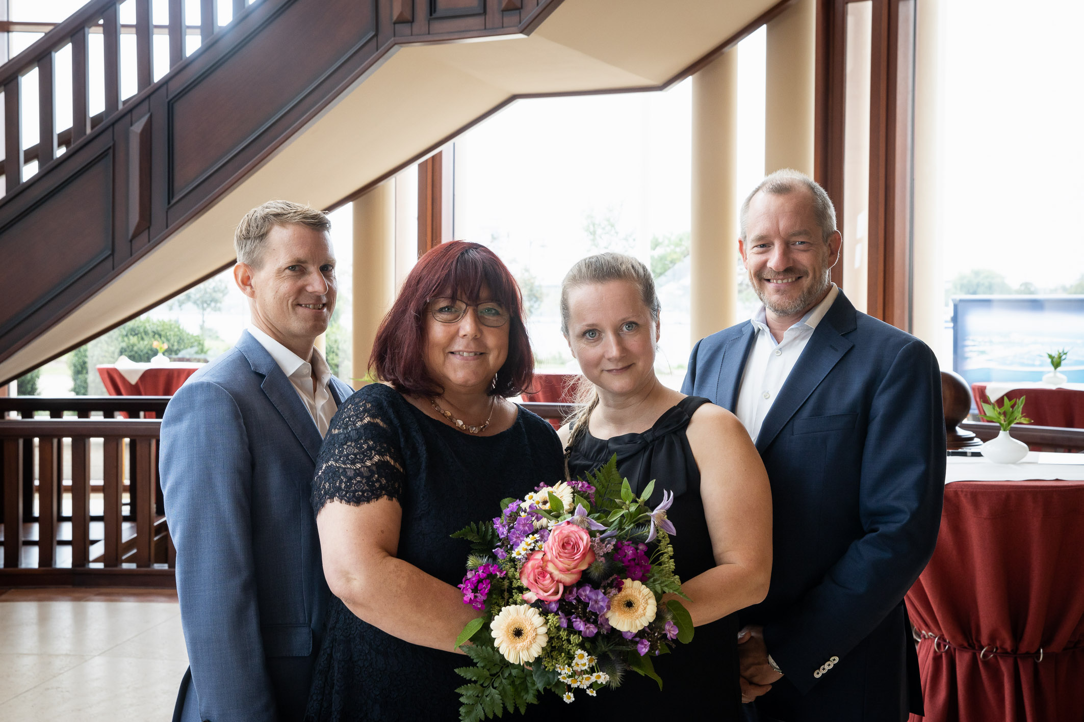Vier festlich gekleidete Personen stehen lächelnd in einem eleganten Raum mit großen Fenstern und Holztreppe. Zwei Frauen in der Mitte halten gemeinsam einen bunten Blumenstrauß. Die Szene wirkt feierlich und herzlich – möglicherweise anlässlich eines bes